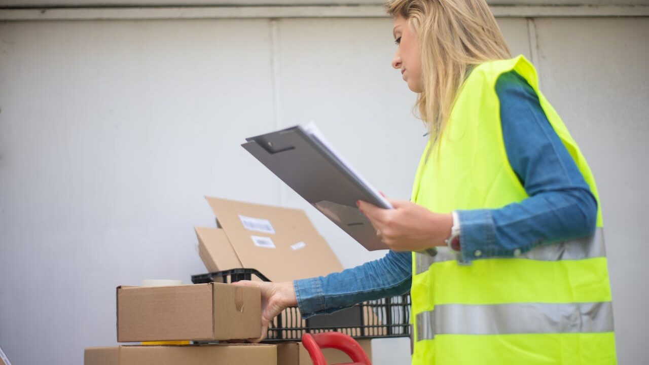 Woman in high-visibility vest handling boxes for delivery service inside a warehouse setting.